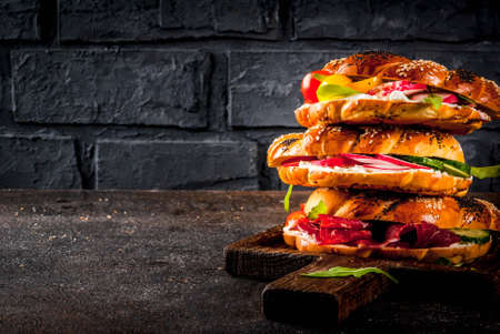 Stack of various homemade bagels sandwiches with sesame and poppy seeds, cream cheese,  ham, radish, arugula, cherry tomatoes, cucumbers, on cutting board. dark concrete background copy space の写真素材