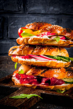 Stack of various homemade bagels sandwiches with sesame and poppy seeds, cream cheese,  ham, radish, arugula, cherry tomatoes, cucumbers, on cutting board. dark concrete background copy space の写真素材