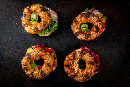 Variety of homemade bagels sandwiches with sesame and poppy seeds, cream cheese,  ham, radish, arugula, cherry tomatoes, cucumbers, dark concrete background copy space aboveの写真素材
