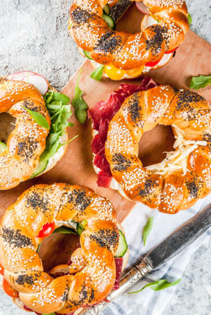 Variety of homemade bagels sandwiches with sesame and poppy seeds, cream cheese,  ham, radish, arugula, cherry tomatoes, cucumbers, white grey textured background copy space top viewの写真素材