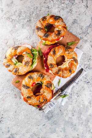 Variety of homemade bagels sandwiches with sesame and poppy seeds, cream cheese,  ham, radish, arugula, cherry tomatoes, cucumbers, white grey textured background copy space top viewの写真素材