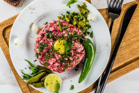 French cuisine, beef steak tartare with raw quail egg yolk, pickled cucumber and onions, fresh herbs, bread, white marble background copy space aboveの写真素材