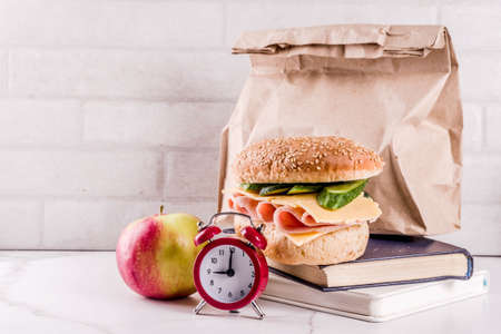 Healthy school food concept, paper bag with lunch, apple, sandwich, books and alarm clock on white kitchen table copy spaceの写真素材