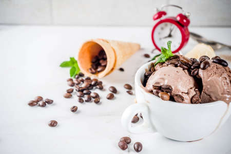 Time for coffee concept, with an alarm clock for hours in the frame. Homemade coffee ice cream, served with coffee beans and mint leaves, with ice cream cones and spoons. White marble background,の写真素材