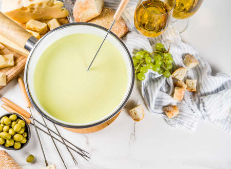 Gourmet Swiss fondue in traditional fondue pot, with forks, various cheeses, olives, bread and grape, white marble background copy spaceの写真素材