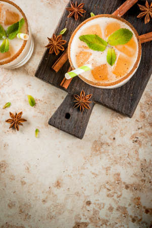 Traditional Indian drink is iced tea or chai masala, with ice cubes from chai, milk and mint leaves. With striped straws, on a wooden board. On light beige stone table. Copy space top viewの写真素材