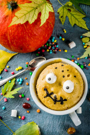 Kids Halloween food idea, funny mug cake decorate like monster face with marshmallow eyes and mouth painted with edible marker, blue background with sweets and autumn leaves, copy spaceの写真素材