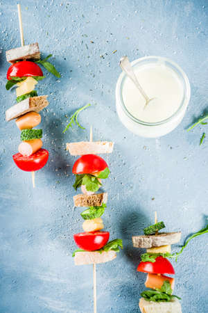 Healthy summer snack idea, salad sandwiches kebab on skewers, with bread slices, tomato, greens, cucumber, cheese, sausages, with yoghurt dipping, on light concrete background copy space top viewの写真素材