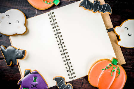 Traditional Halloween baked pastry, funny cookies for children's treat - ghost, pumpkins, black cat, bats, witch house. On old wooden table, top view copy space, with notebook for recipesの写真素材