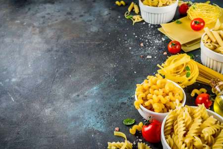 Italian food concept, various raw pasta assortment - spaghetti, lasagna, fusilli, tagliatelle, penne, tortellini, ravioli, with tomatoes and basil leaves dark background copy space top viewの写真素材