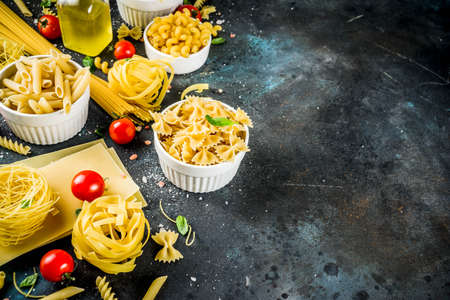 Italian food concept, various raw pasta assortment - spaghetti, lasagna, fusilli, tagliatelle, penne, tortellini, ravioli, with tomatoes and basil leaves dark background copy space top viewの写真素材