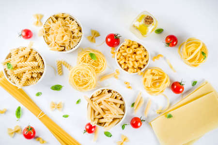 Italian food concept, various raw pasta assortment - spaghetti, lasagna, fusilli, tagliatelle, penne, tortellini, ravioli, with tomatoes and basil leaves white background copy space top viewの写真素材