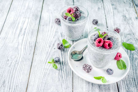 Summer healthy breakfast dessert in glass, overnight vegan chia pudding with non-dairy milk, frozen raspberry, blackberry and blueberry, on light blue and wooden background copy spaceの写真素材