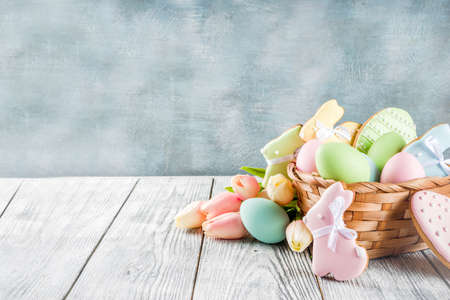 Easter greeting card background with pastel colored eggs and homemade cookies shaped in eggs and bunnies rabbits. With a basket, tulips, rustic wooden table, copy spaceの写真素材