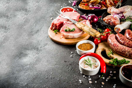 Various raw meat ready for grill and bbq, with vegetables, greens, sauces kitchen grilling utensils. Chicken legs, pork steaks, sausages, beef ribs with herbs, black concrete background copy spaceの写真素材