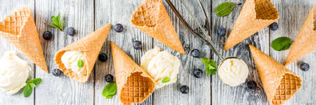 White vanilla ice cream balls with ice cream cones, spoon, fresh blueberry berries and mint leaves, rustic wooden background copy space bannerの写真素材
