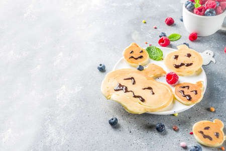 Halloween kids food treat. Funny pancakes - ghosts, jack o Lantern pumpkins, spider webs. Breakfast snack for child's Halloween party, with berries & candy,  gray stone table top view copy spaceの写真素材