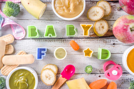 Infant baby food. Bowls with vegetable fruit puree, green, orange, yellow colors - broccoli, carrots, banana, apple. With baby accessories and toys wooden white background Flat lay top viewの写真素材