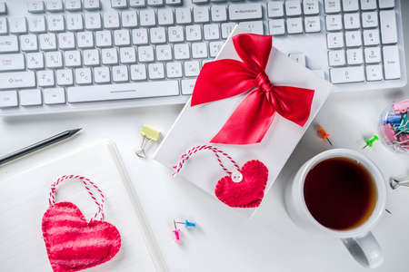 Valentine day concept. Secret Valentine office game idea. February 14 holiday background with gift box, textile hearts and office supplies, white keyboard and notepad. White table background copy spaceの写真素材