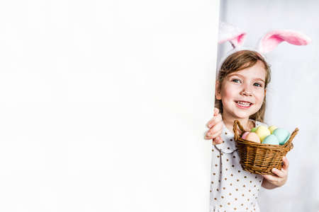 Happy cute child girl in a rabbit costume with hare ears with a basket of painted eggs peeks out from behind the wall.の写真素材