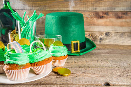 Food for kids celebrating St. Patrick day party. Green leprechaun hat with green sweets - sweets, drink, rainbow cupcakes and golden chocolate coins. On a wooden table.の写真素材