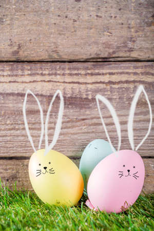 Colorful pastel Easter eggs with drawn bunny ears on green and wooden garden terrace. Funny spring...