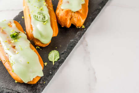 Traditional French dessert. Mojito Eclairs with lime zest and mint leaves, on dark blue background,...