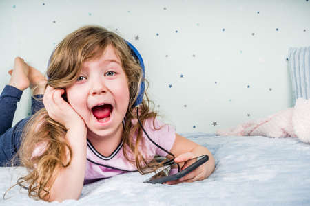Kids Gaming video games concept. Toddler or young girl, playing game with joystick, smart-phone, VR glasses and headphones, enjoying games, sitting on sofa in living room at home.の写真素材