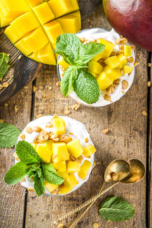 Summer breakfast layered dessert with mango and granola. Mango parfaits in portioned jars, rustic wood background copy spaceの写真素材