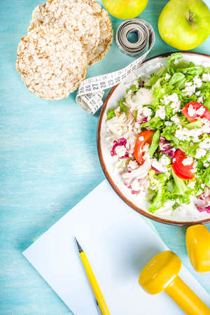 Healthy eating, slimming. dieting and weigh loss concept background. Fresh greenery summer salad with fruit, measuring tape, dumbbells and diet plan dairy notepad, flat lay above copy spaceの写真素材