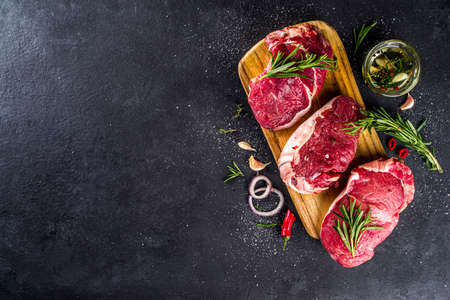 Raw meat, beef steak on cutting board with rosemary and spices, black background, top view copy...