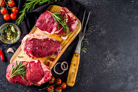 Raw meat, beef steak on cutting board with rosemary and spices, black background, top view copy...