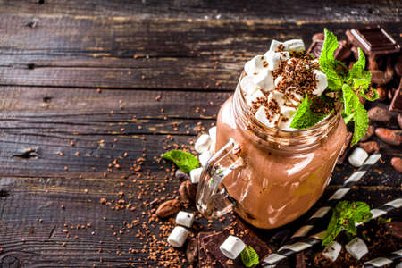 Chocolate milk shake with sauce, marshmallows and grated chocolate, decorated with mint. On old rustic wooden table with a lot of chocolate pieces, cocoa beans and chocolate shavingsの写真素材