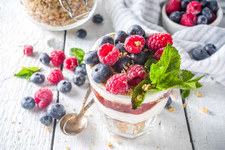 Yogurt parfafait with granola and berries. Sweet and healhty breakfast dessert in glass with granola, Yogurt, blueberries and raspberries. White wooden background top viewの写真素材