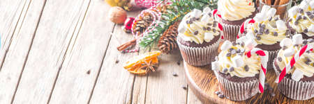 Christmas cupcakes. Xmas hot chocolate cupcake with peppermint candy cane, chocolate drops and marshmallow. Wooden background with Christmas decor and fir tree branches. Sweet Christmas baking /の写真素材