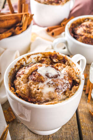 Cinnabon cake in mug. Fast simple microwave dessert idea, background for recipe. Cinnamon roll mugcake, with sugar and cream cheese topping, in different mugs, with cinnamon sticks on wooden backgroundの写真素材
