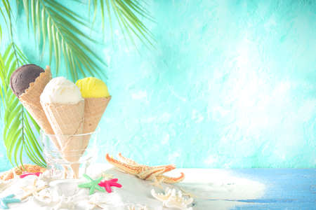 Summer tropical sea holiday vacation concept. three ice cream waffle cones with different flavors of ice-cream, on sand with seashells and starfish, light blue wooden backgroundの写真素材