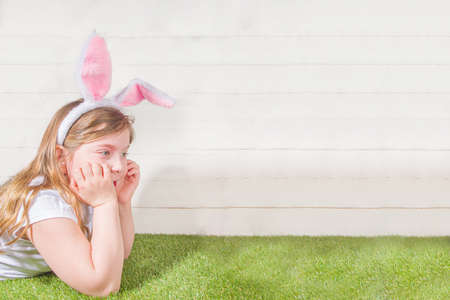 Cute little child girl wearing bunny ears on Easter holiday. Girl hunts for Easter eggs in the garden, on Spring Grass and White Wooden Background copy spaceの写真素材