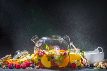 Fruit and berry tea in teapot. Hot drink with lemon, mint, blueberry, ginger, orange, apple. Hot flavored steam beverage on dark blue background copy spaceの写真素材