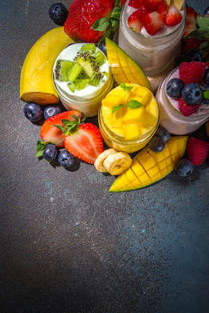 Set of various fruit and berry sweet yogurts in glass jars. Variety healthy Breakfast yoghurts with blueberry, strawberry, mango, kiwi, raspberry, with fresh fruits and berries, dark backgroundの写真素材
