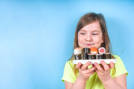 Cute little girl enjoys eating sushi. Cheerful funny caucasian blond preschool child girl with various sushi roll and chopsticks. On bright blue backgroundの写真素材