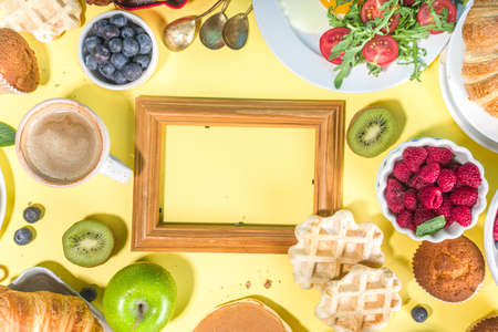 Various traditional breakfast food - fried eggs with bacon, muesli, oats, waffles, pancakes, burger, croissants, fruit berry, coffee, tea and orange juice, yellow table background copy space top viewの写真素材