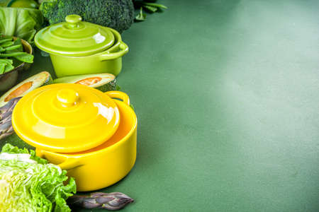 Various organic green spring vegetable ingredients with empty colorful portioned cooking pots on dark green kitchen table, top view. Diet healthy soup, stew food preparation conceptの写真素材