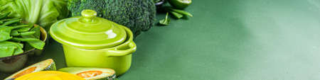 Various organic green spring vegetable ingredients with empty colorful portioned cooking pots on dark green kitchen table, top view. Diet healthy soup, stew food preparation conceptの写真素材