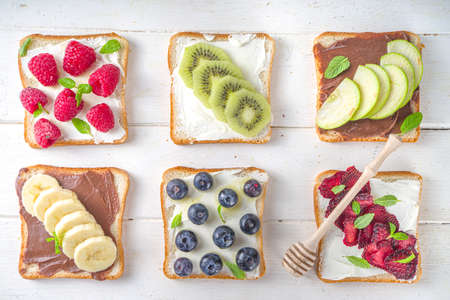 Healthy fruit and berry sandwiches. Tasty diet toast bread with cream cheese and banana, apple, raspberry, blueberry, strawberry, on white wooden background top view copy spaceの写真素材