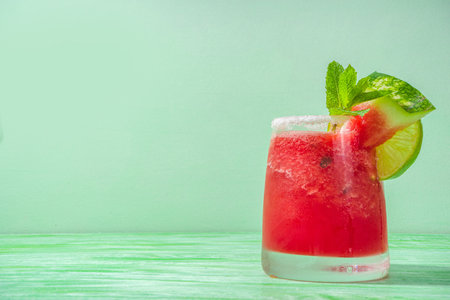 Cold summer cocktail, watermelon margaritas or mojito with watermelon and lime slices, crushed ice and mint. Seasonal refreshing drink, on bright green sun lighted backgroundの写真素材
