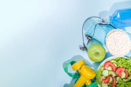 Sport healthy lifestyle, diet concept. Slimming weight loss fitness background with measuring tape, colored dumbbells, salad, cereal crisp bread, sports water bottle on Blue background copy spaceの写真素材