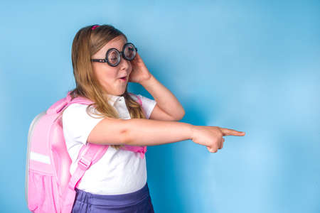 Back to school concept. School invitation, advertisement banner. Cute primary student schoolgirl in white blue classic uniform, with books backpack, funny glasses. Colorful blue background copy spaceの写真素材