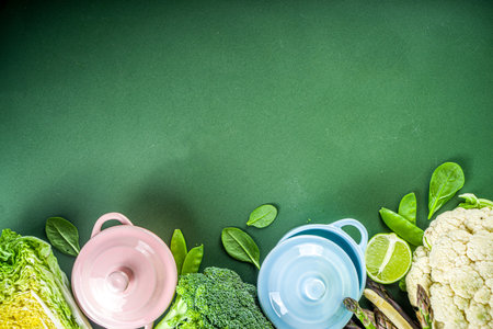 Various organic green spring vegetable ingredients with empty colorful portioned cooking pots on dark green kitchen table, top view. Diet healthy soup, stew food preparation conceptの写真素材
