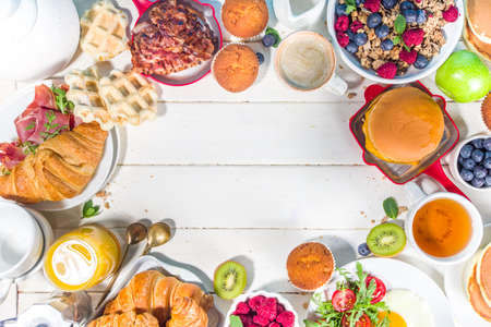 Various traditional breakfast food - fried eggs with bacon, muesli, oats, waffles, pancakes, burger, croissants, fruit berry, coffee, tea and orange juice, white table background copy space top viewの写真素材